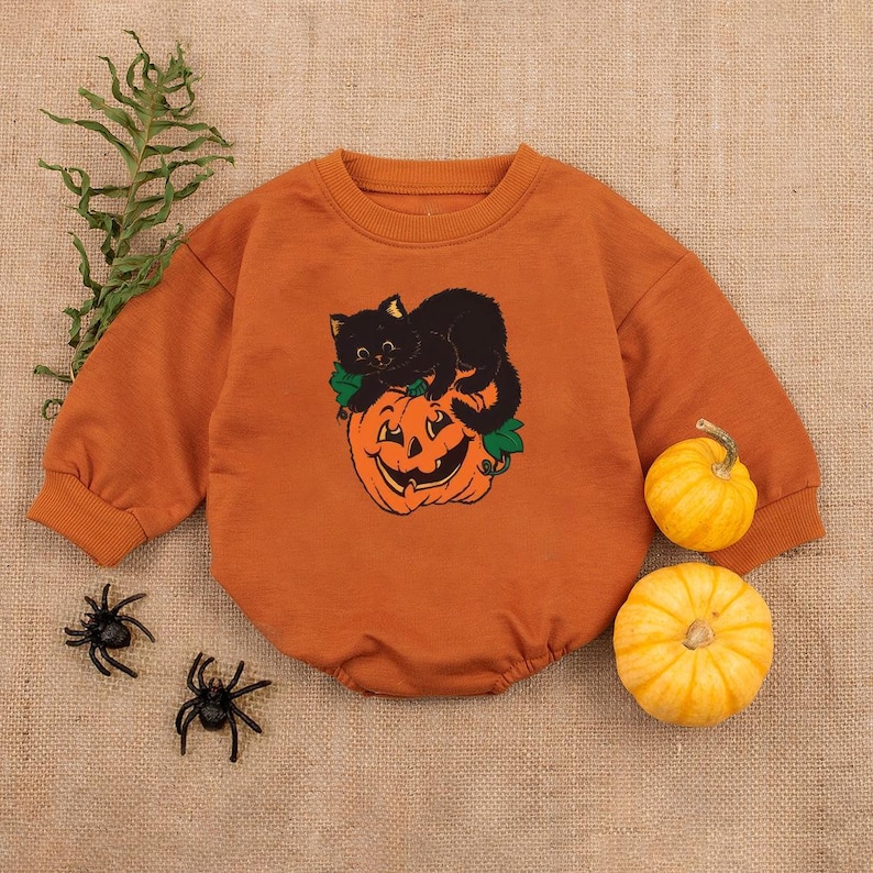 May include: An orange baby romper with a black cat and jack-o'-lantern graphic. The long-sleeved outfit has a crew neck and is decorated with a smiling pumpkin and a black cat. Two small pumpkins and plastic spiders are also in the image.