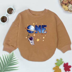 May include: A brown long-sleeved sweatshirt with a graphic of an astronaut holding a planet with the word "ONE" printed on it. The graphic also includes stars and a rocket.