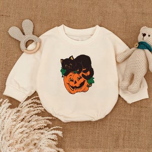 May include: Cream-colored baby onesie featuring a black cat resting on an orange jack-o'-lantern with green leaves. The onesie has long sleeves and a round neckline. A beige bunny-shaped teether and a stuffed bear are also in the image.