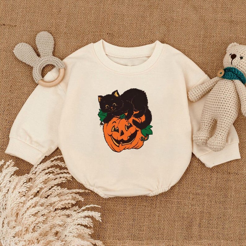 May include: Cream-colored baby onesie featuring a black cat resting on an orange jack-o'-lantern with green leaves. The onesie has long sleeves and a round neckline. A beige bunny-shaped teether and a stuffed bear are also in the image.
