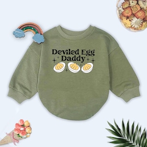 May include: Olive green sweatshirt with the text "Deviled Egg Daddy" and three deviled egg illustrations. The sweatshirt has a crew neck and long sleeves. A colorful rainbow decoration is on the top left.