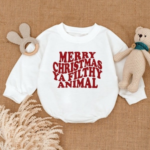 May include: White baby onesie with the text "MERRY CHRISTMAS YA FILTHY ANIMAL" in red lettering. A wooden bunny teether and a small teddy bear are next to the onesie on a brown surface. The onesie has long sleeves.