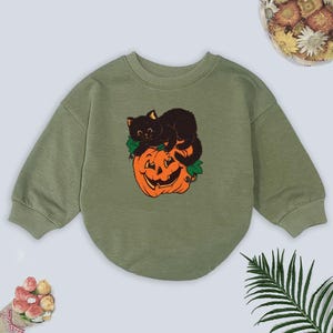 May include: Olive green sweatshirt featuring a Halloween-themed graphic. The graphic depicts a black cat resting on top of a carved orange pumpkin. The sweatshirt has long sleeves and a rounded neckline, perfect for the fall season.