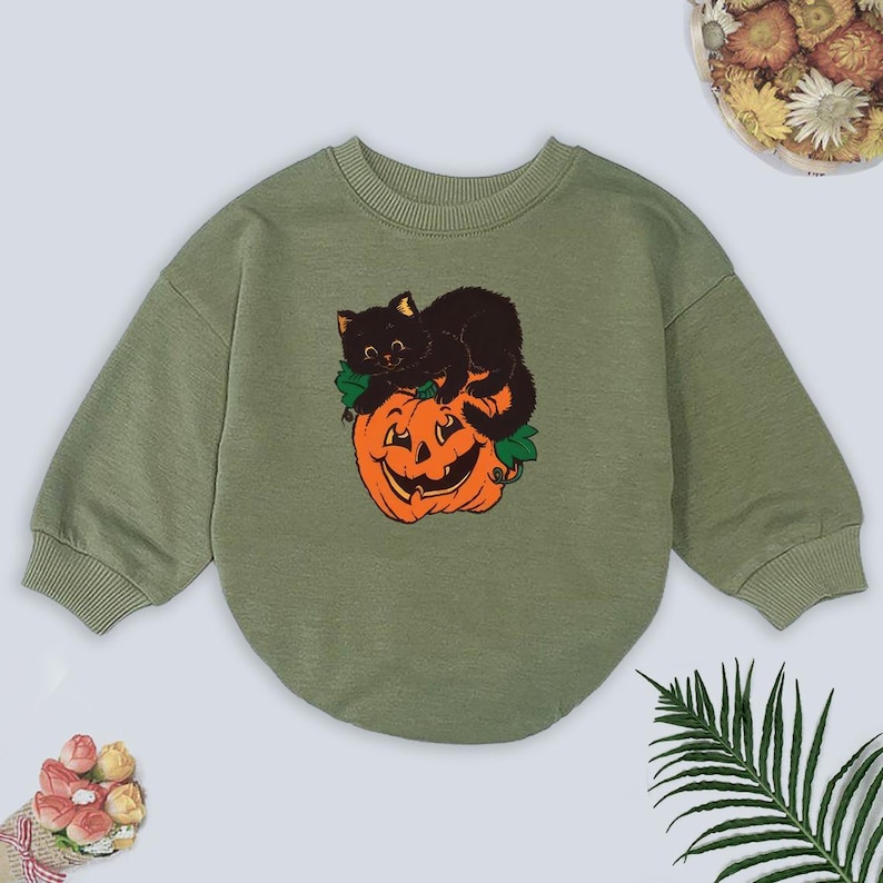 May include: Olive green sweatshirt featuring a Halloween-themed graphic. The graphic depicts a black cat resting on top of a carved orange pumpkin. The sweatshirt has long sleeves and a rounded neckline, perfect for the fall season.