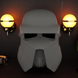 Könnte beinhalten: Ein grauer Stormtrooper-Helm ist der Mittelpunkt dieses Bildes, vor einem dunklen Hintergrund. Zwei Wandleuchten mit kugelförmigen, gelb leuchtenden Lichtern flankieren den Helm. Eine dekorative Illustration eines Roboterkopfes ist über der linken Leuchte zu sehen.