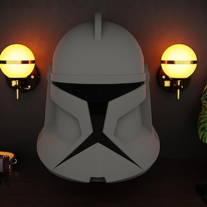 May include: A grey and black Clone Trooper helmet from Star Wars is displayed on a dark wooden surface. Two wall sconces with spherical, glowing yellow lights flank the helmet. A decorative graphic is mounted on the wall above the sconces.