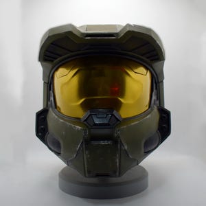 May include: An olive green helmet with a gold visor, resembling a futuristic military design. The helmet has a detailed, textured surface and is mounted on a grey base. The visor reflects light, adding a metallic sheen.