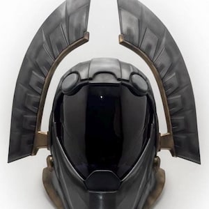 Puede incluir: Un casco futurista en tonos grises y bronce. El casco presenta una visera oscura y está flanqueado por dos grandes estructuras en forma de alas. El diseño sugiere un tema de ciencia ficción o cosplay.