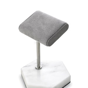 May include: A gray suede watch display stand with a stainless steel post and a white marble hexagonal base. The stand is designed to hold a single watch. The base has a black rubber bottom.