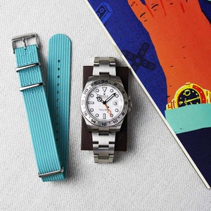 K&ouml;nnte beinhalten: Eine wei&szlig;e Rolex-Uhr mit schwarzer L&uuml;nette und wei&szlig;em Zifferblatt. Die Uhr hat ein Datumsfenster bei 3 Uhr und einen schwarzen Sekundenzeiger. Die Uhr ist an einem silbernen Metallband. Die Uhr liegt auf einer braunen Oberfl&auml;che mit blauem und rotem Hintergrund.