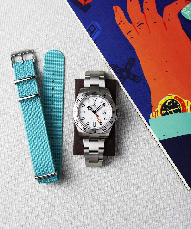 K&ouml;nnte beinhalten: Eine wei&szlig;e Rolex-Uhr mit schwarzer L&uuml;nette und wei&szlig;em Zifferblatt. Die Uhr hat ein Datumsfenster bei 3 Uhr und einen schwarzen Sekundenzeiger. Die Uhr ist an einem silbernen Metallband. Die Uhr liegt auf einer braunen Oberfl&auml;che mit blauem und rotem Hintergrund.