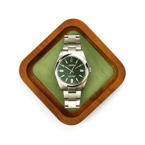 Puede incluir: Un reloj de pulsera de acero inoxidable con esfera verde y agujas plateadas descansa en una bandeja de madera con forro de fieltro verde. El reloj tiene una correa plateada y una esfera redonda. La bandeja es cuadrada con esquinas redondeadas.