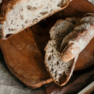 May include: A close-up of a loaf of sourdough bread sliced on a wooden cutting board. The bread has a light brown crust and a soft, airy interior. The bread is sliced into two pieces, one of which is partially eaten.