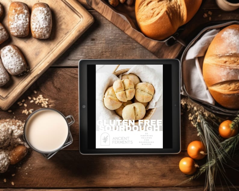 May include: A digital tablet displaying a book cover with the title "Gluten Free Sourdough" and the text "20 Inspiring Recipes for Sourdough Bakers" by Ancient Ferments. The cover features a basket of freshly baked sourdough rolls.