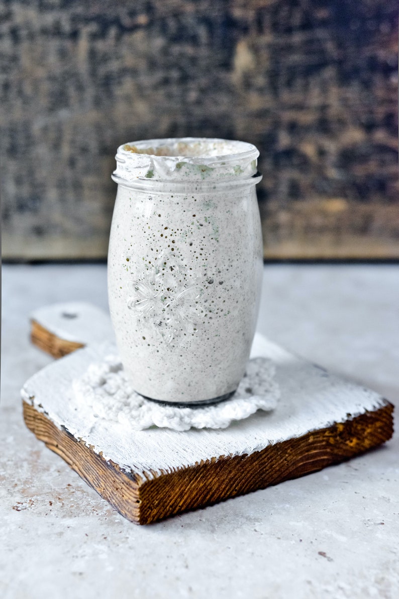 May include: A glass jar filled with a white and gray mixture, possibly sourdough starter, sitting on a white wooden cutting board with a white crocheted doily.