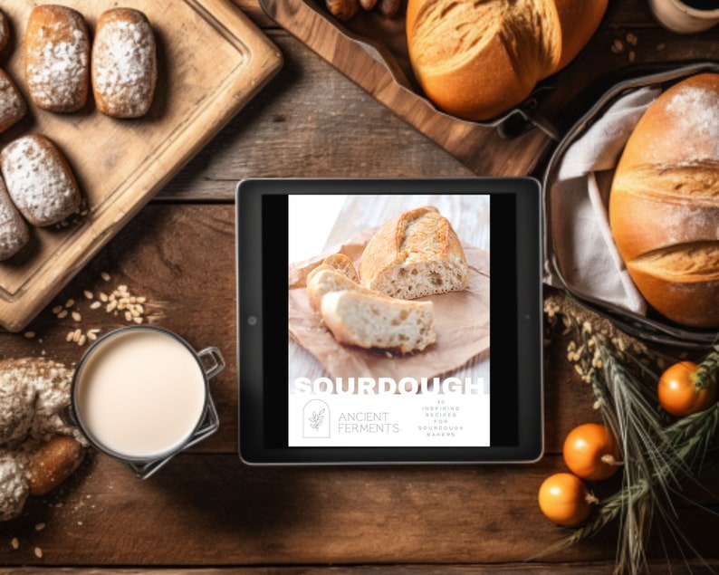 May include: Overhead shot of a wooden table with various baked goods. A tablet displays the word "SOURDOUGH" with the text "20 Inspiring Recipes for Sourdough Bakers." There are loaves of bread, rolls, and a glass of milk.