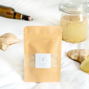 May include: A brown paper pouch with a white label that says "Ancient Ferments" and a small illustration of a plant. The pouch contains a white powder. There are pieces of ginger on a white surface.