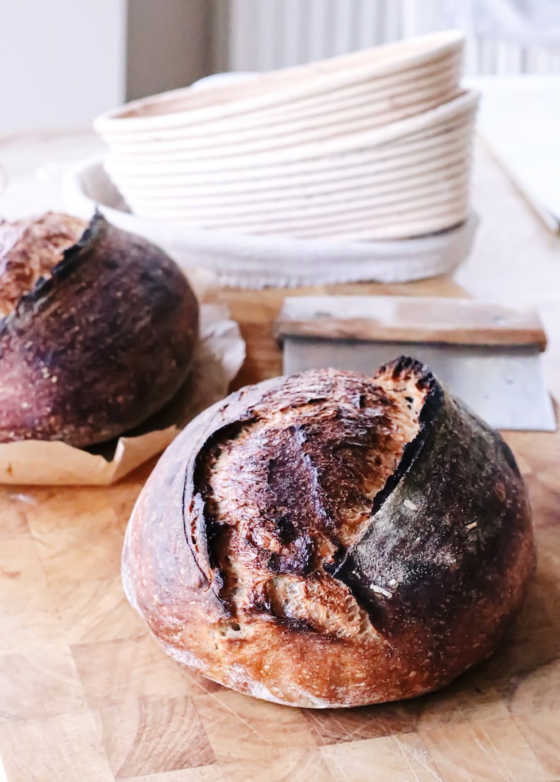 May include: Two loaves of freshly baked sourdough bread on a wooden cutting board. The bread has a dark brown crust and a light brown interior.