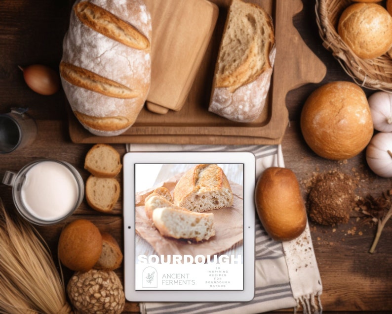 May include: Overhead shot of various breads, including a large loaf and rolls, arranged on a wooden surface. A tablet displays the word "SOURDOUGH" with text below. Ingredients like flour, garlic, and an onion are also present, suggesting a baking theme.
