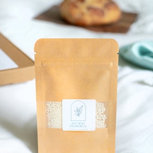 May include: A brown paper pouch with a clear window showing a white sourdough starter inside. The pouch has a white label with the text "Ancient Ferments" and "Sourdough Starter".