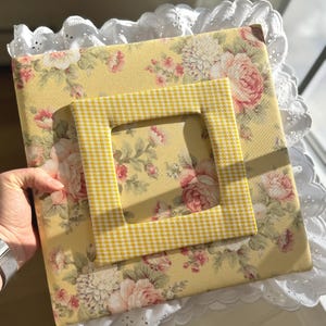 Könnte beinhalten: Ein gelbes Fotoalbum mit Blumenmuster und einem gelb-weiß karierten Rahmen. Das Album ist mit einer weißen Spitzenborte verziert. Das Blumenmuster umfasst rosa und weiße Rosen.