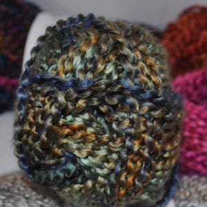 May include: A round ball of thick, hand-spun yarn featuring a variegated colour palette of deep forest green, metallic bronze, and navy blue. The yarn has a high-sheen, twisted texture with visible individual fibres woven into a dense, chunky structure.