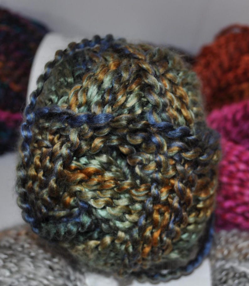 May include: A round ball of thick, hand-spun yarn featuring a variegated colour palette of deep forest green, metallic bronze, and navy blue. The yarn has a high-sheen, twisted texture with visible individual fibres woven into a dense, chunky structure.