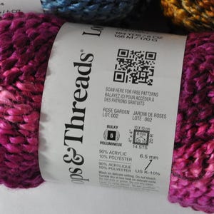 May include: A skein of bulky weight yarn in a variegated magenta and dark pink colourway. The white paper label features the brand name Loops & Threads, a QR code, and technical specifications including 90% acrylic and 10% polyester content.