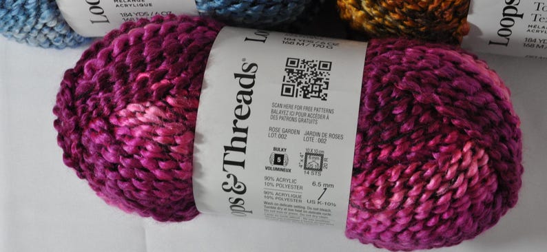 May include: A skein of bulky weight yarn in a variegated magenta and dark pink colourway. The white paper label features the brand name Loops & Threads, a QR code, and technical specifications including 90% acrylic and 10% polyester content.