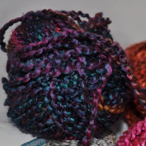 May include: A round ball of thick, multi-coloured yarn featuring a blend of deep purple, dark blue, and hints of copper and teal. The yarn has a twisted, textured appearance with a slight sheen, showing a continuous strand wrapped into a sphere.