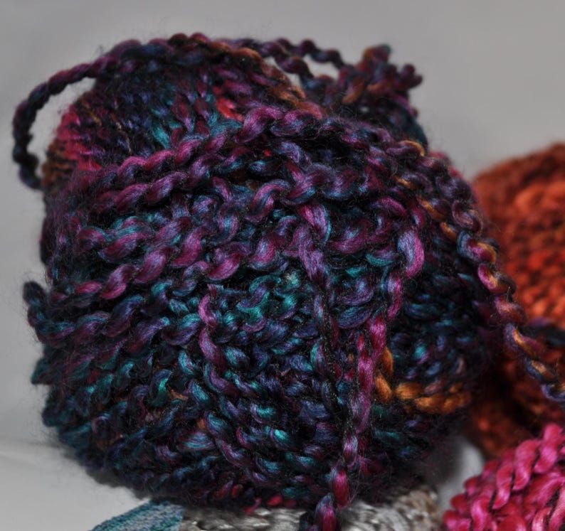 May include: A round ball of thick, multi-coloured yarn featuring a blend of deep purple, dark blue, and hints of copper and teal. The yarn has a twisted, textured appearance with a slight sheen, showing a continuous strand wrapped into a sphere.