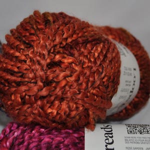 May include: A ball of rust-coloured yarn with a thick, twisted texture. A white paper label wraps around the centre, featuring the text Loops & Threads, a QR code, and the words Rose Garden. The yarn has a slight sheen and a chunky, braided appearance.