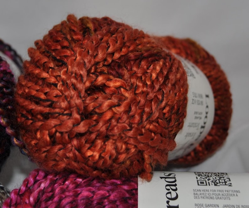 May include: A ball of rust-coloured yarn with a thick, twisted texture. A white paper label wraps around the centre, featuring the text Loops & Threads, a QR code, and the words Rose Garden. The yarn has a slight sheen and a chunky, braided appearance.