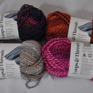 May include: Four skeins of Loops & Threads Textured Twist acrylic blend yarn. The colors include a dark purple and black mix, a burnt orange and brown blend, a grey and tan variegated mix, and a vibrant magenta and pink variegated yarn.