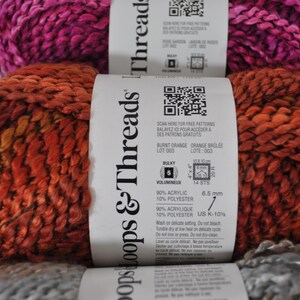 May include: Three skeins of Loops & Threads bulky yarn stacked vertically. The top skein is magenta, the middle is burnt orange, and the bottom is grey. Each skein has a white paper label with a QR code, fibre content of 90% acrylic and 10% polyester.