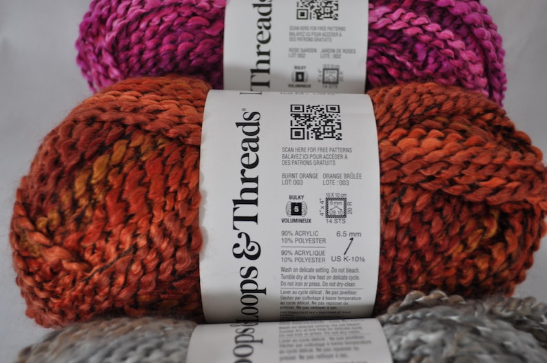 May include: Three skeins of Loops & Threads bulky yarn stacked vertically. The top skein is magenta, the middle is burnt orange, and the bottom is grey. Each skein has a white paper label with a QR code, fibre content of 90% acrylic and 10% polyester.