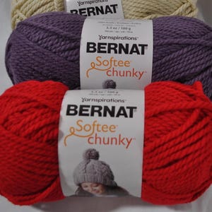 Puede incluir: Tres ovillos de lana Bernat Softee Chunky apilados verticalmente. El ovillo superior es crema, el del medio es morado y el inferior es rojo. Cada ovillo tiene una etiqueta de papel blanco con la marca, el nombre y el peso de 100 g.