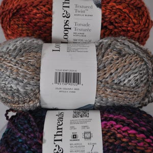 May include: Three skeins of Loops & Threads Textured Twist acrylic blend yarn stacked vertically. The top skein is burnt orange, the middle is a mix of grey and tan, and the bottom is a dark blend of purple, blue, and black. Each has a white label.