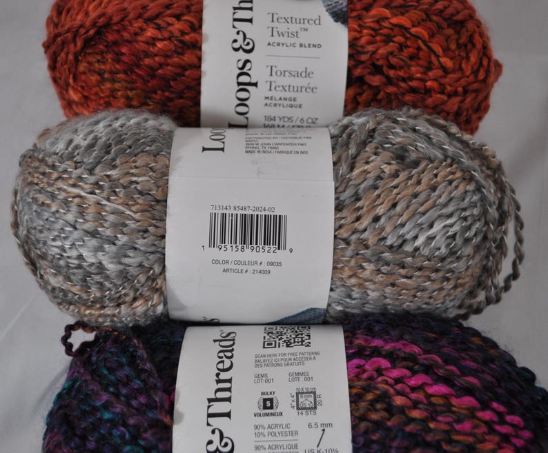 May include: Three skeins of Loops & Threads Textured Twist acrylic blend yarn stacked vertically. The top skein is burnt orange, the middle is a mix of grey and tan, and the bottom is a dark blend of purple, blue, and black. Each has a white label.