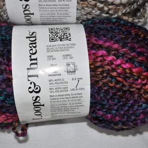 May include: Two skeins of Loops & Threads yarn featuring a variegated colour palette of deep purple, bright magenta, and dark blue. The white paper label displays a QR code, 90% acrylic and 10% polyester content, and a 6.5 mm needle size requirement.