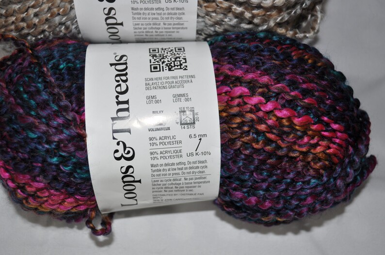 May include: Two skeins of Loops & Threads yarn featuring a variegated colour palette of deep purple, bright magenta, and dark blue. The white paper label displays a QR code, 90% acrylic and 10% polyester content, and a 6.5 mm needle size requirement.