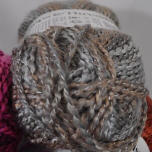 May include: A skein of yarn featuring a variegated blend of grey, silver, and light brown fibres. The yarn has a thick, twisted texture and is wrapped with a white paper label that partially obscures the top and bottom of the round ball.