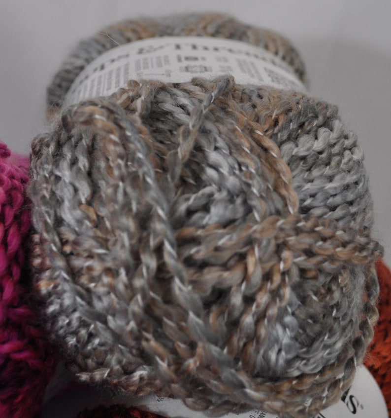 May include: A skein of yarn featuring a variegated blend of grey, silver, and light brown fibres. The yarn has a thick, twisted texture and is wrapped with a white paper label that partially obscures the top and bottom of the round ball.