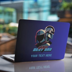 Op de afbeelding: Laptop met een donkerblauwe skin met een gestileerde illustratie van twee helmen in teal en roze. De tekst luidt "GALAXY'S BEST DAD" en "YOUR TEXT HERE". De laptop staat op een tafel met een vage achtergrond.