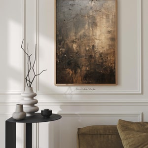 May include: Abstract painting in shades of brown, gray, and gold with a textured surface. The painting is framed in a thin, wooden frame and is hanging on a white wall.