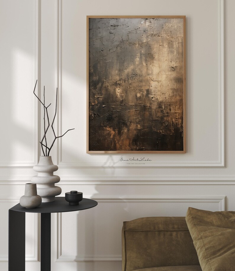 May include: Abstract painting in shades of brown, gray, and gold with a textured surface. The painting is framed in a thin, wooden frame and is hanging on a white wall.