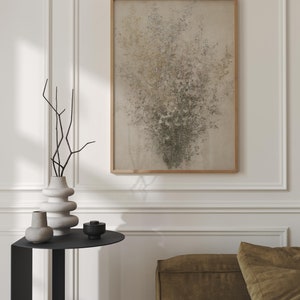 May include: A black side table with a round top and a vase with dried branches, a smaller vase, and a small black bowl. A framed abstract painting with a beige background and green and brown accents hangs on the wall behind the table. A brown couch is in the background.