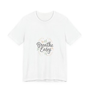 Op de afbeelding: Wit t-shirt met de tekst "Breathe Easy" in een sierlijk lettertype, omringd door een ontwerp van bladeren en stippen. Een casual kledingstuk voor dagelijks gebruik.