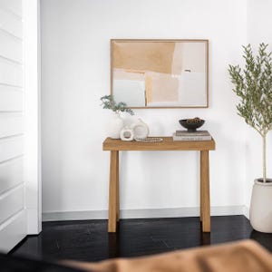 Puede incluir: Una mesa consola rectangular de madera con cuatro patas rectas, sobre la que hay un jarrón blanco, dos pequeñas esculturas de cerámica, un collar de cuentas, un cuenco negro y un libro. Sobre la mesa cuelga un cuadro abstracto enmarcado.