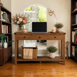 Puede incluir: Una mesa consola rectangular de madera con tres cajones, pomos negros y un estante inferior. La mesa sostiene un portátil, un jarrón de cristal con flores rosas, una vela y una planta en maceta. Una cesta tejida con una manta beige descansa en el estante.